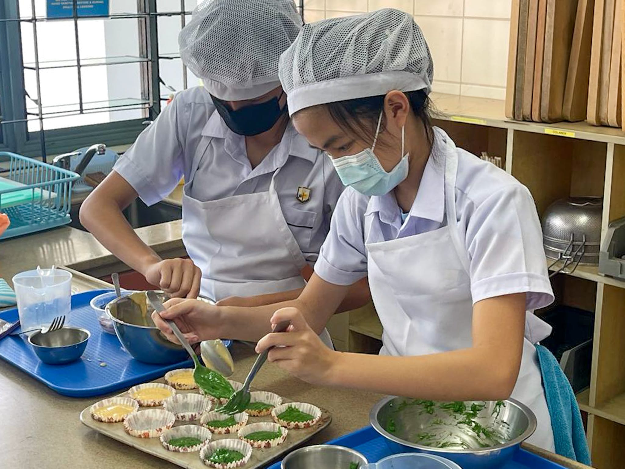 Two masked students in aprons and hairnets spoon green and yellow batter into cupcake liners in a kitchen classroom.